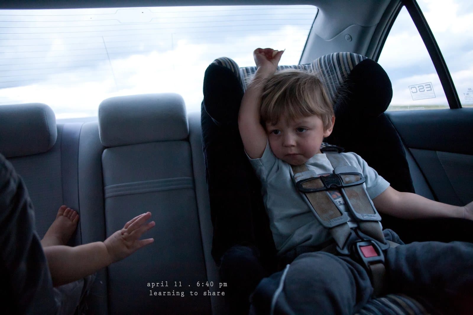 A toddler in a car seat raises his arm while a baby's hand reaches over, captioned learning to share.
