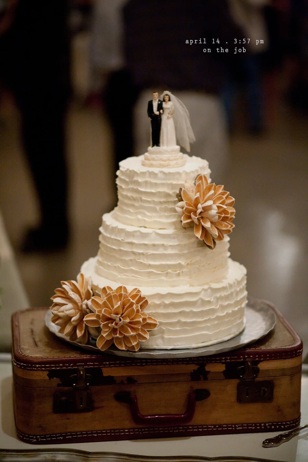 A ruffled white wedding cake with a bride and groom topper sits on a vintage suitcase.