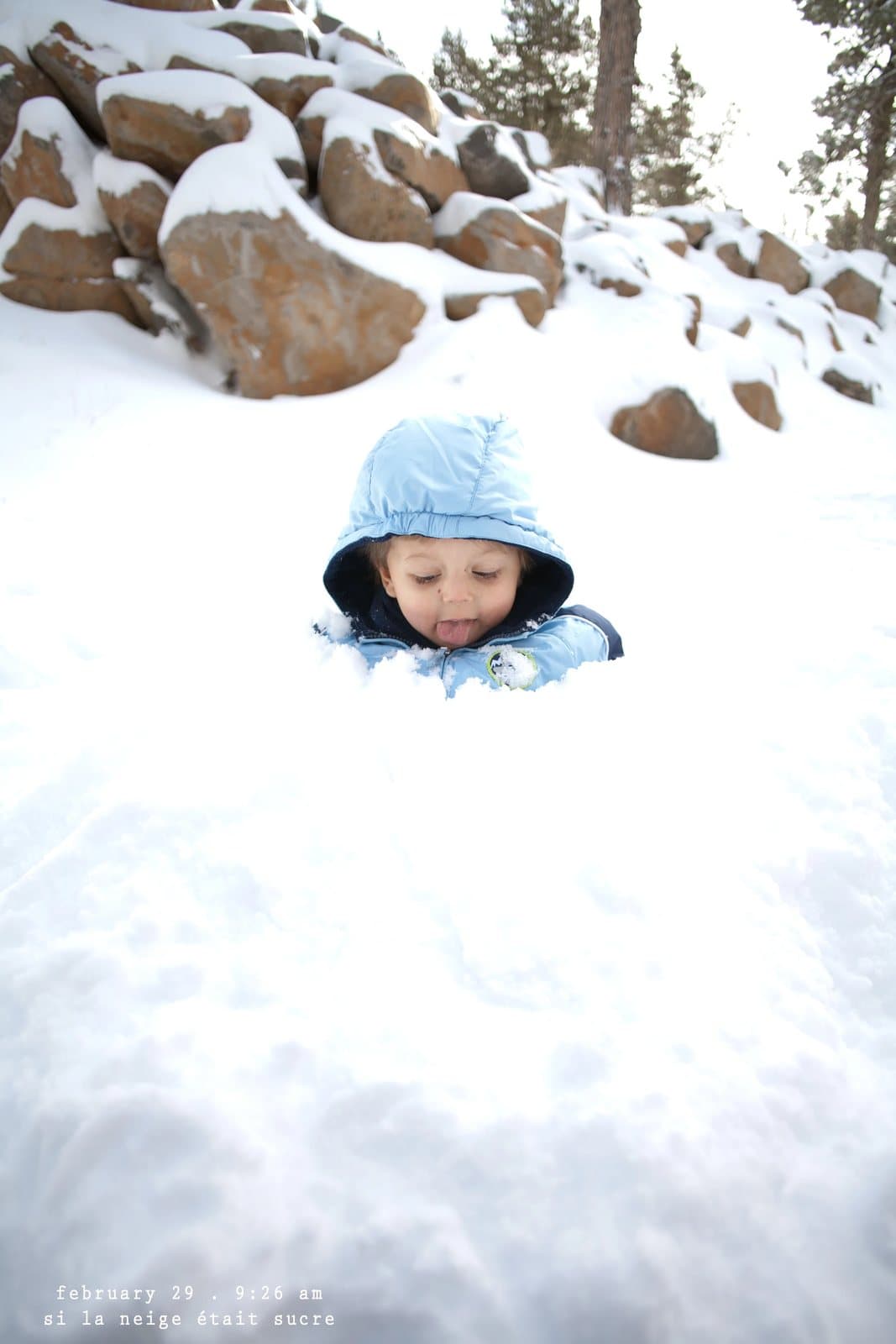 A toddler in a blue hood peeks out of deep snow, captioned si la neige etait sucre.