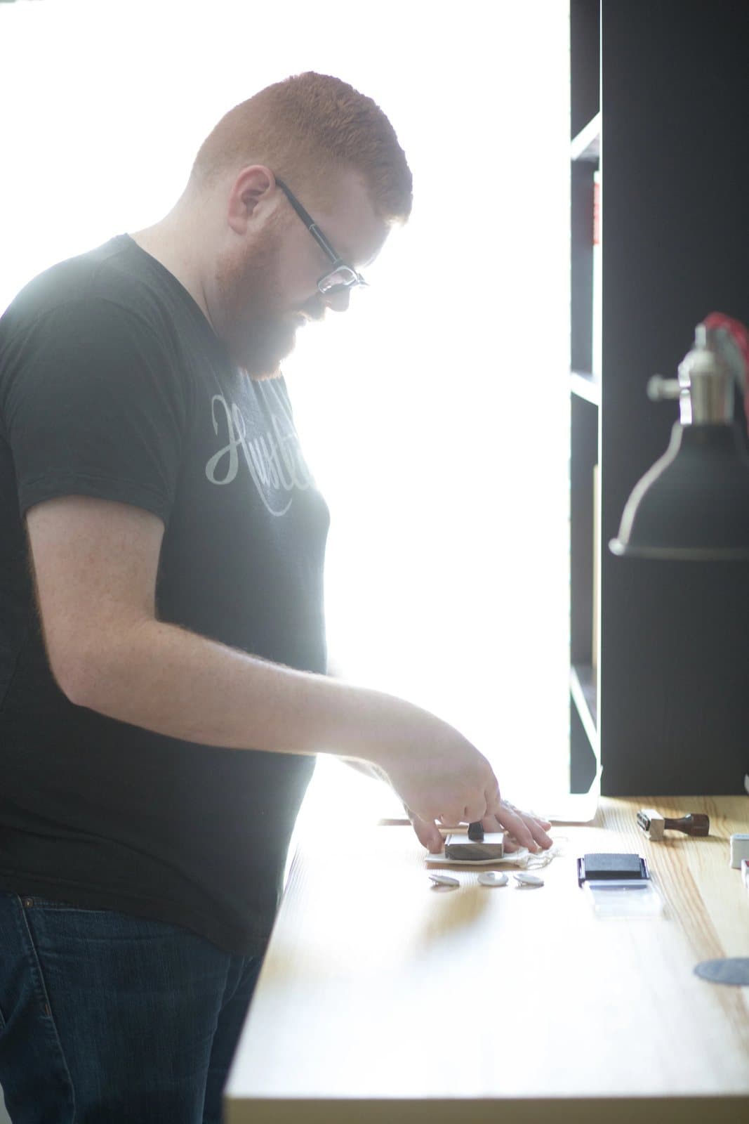 Kyle leans over his bright studio desk, arranging stamps and buttons in window light.