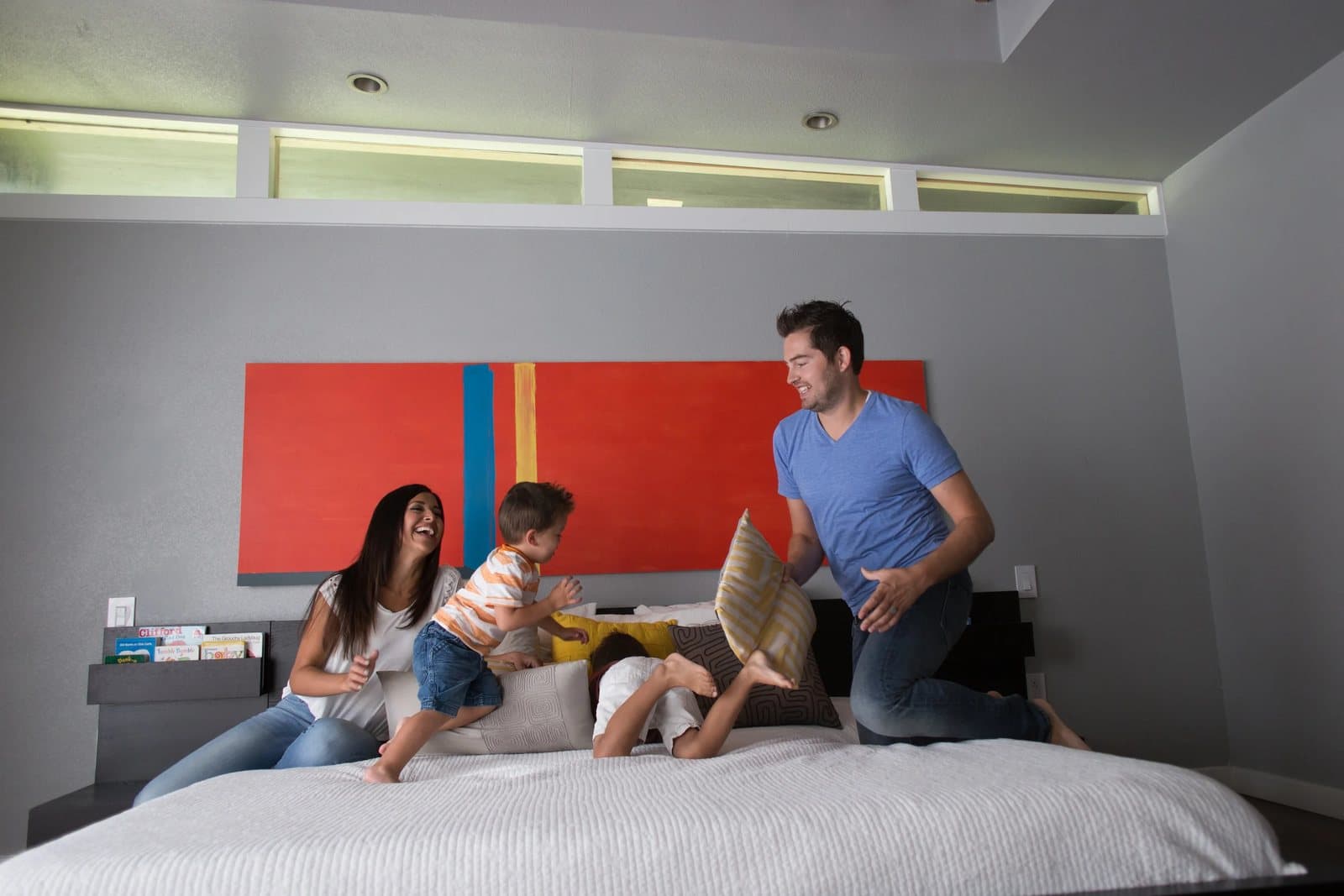The family has a pillow fight on the big bed beneath a red abstract painting.