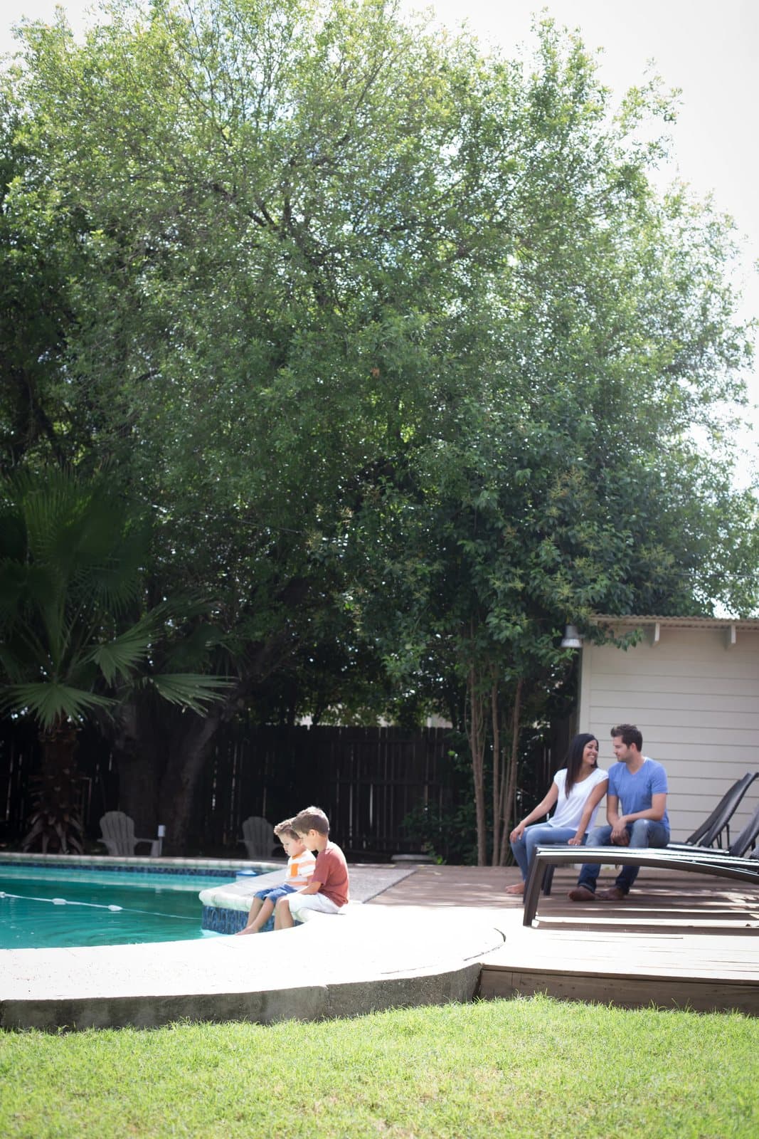 The family relaxes by the backyard pool beneath a large shade tree.